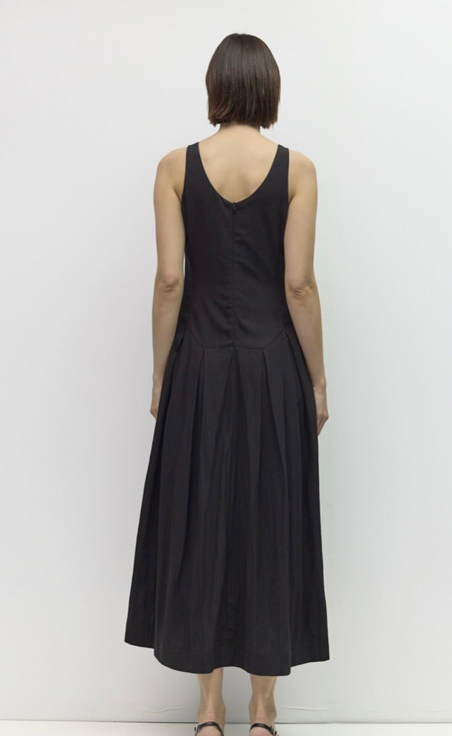 A woman viewed from the back wearing a sleeveless black midi dress with a V-neckline and gathered skirt, standing against a plain light gray wall.