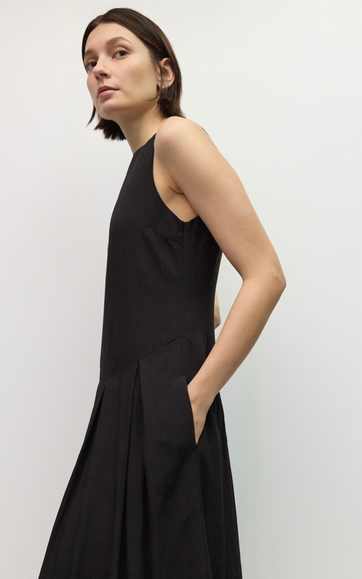 A woman wearing a sleeveless black midi dress with side pockets, photographed against a plain light gray studio background.