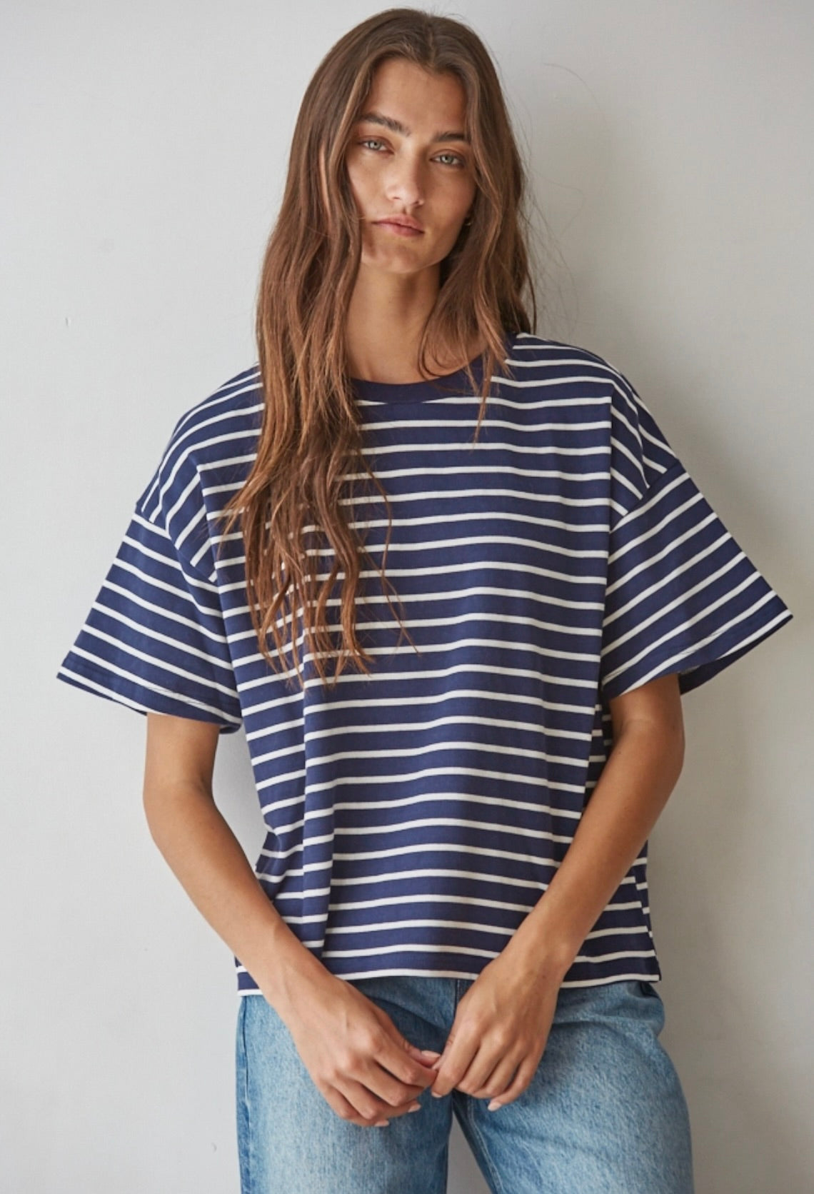 A woman wearing an oversized short-sleeve t-shirt with navy blue and white horizontal stripes, paired with light blue jeans, standing against a plain light gray wall.