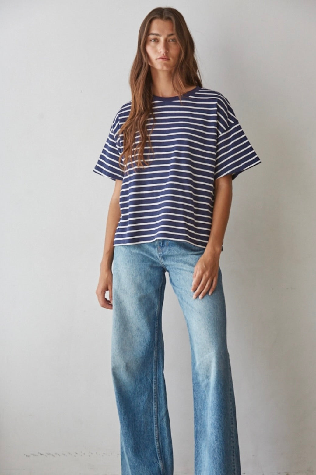 A woman modeling a navy blue and white horizontally striped short-sleeve t-shirt with light-wash blue wide-leg jeans, standing against a plain off-white wall.