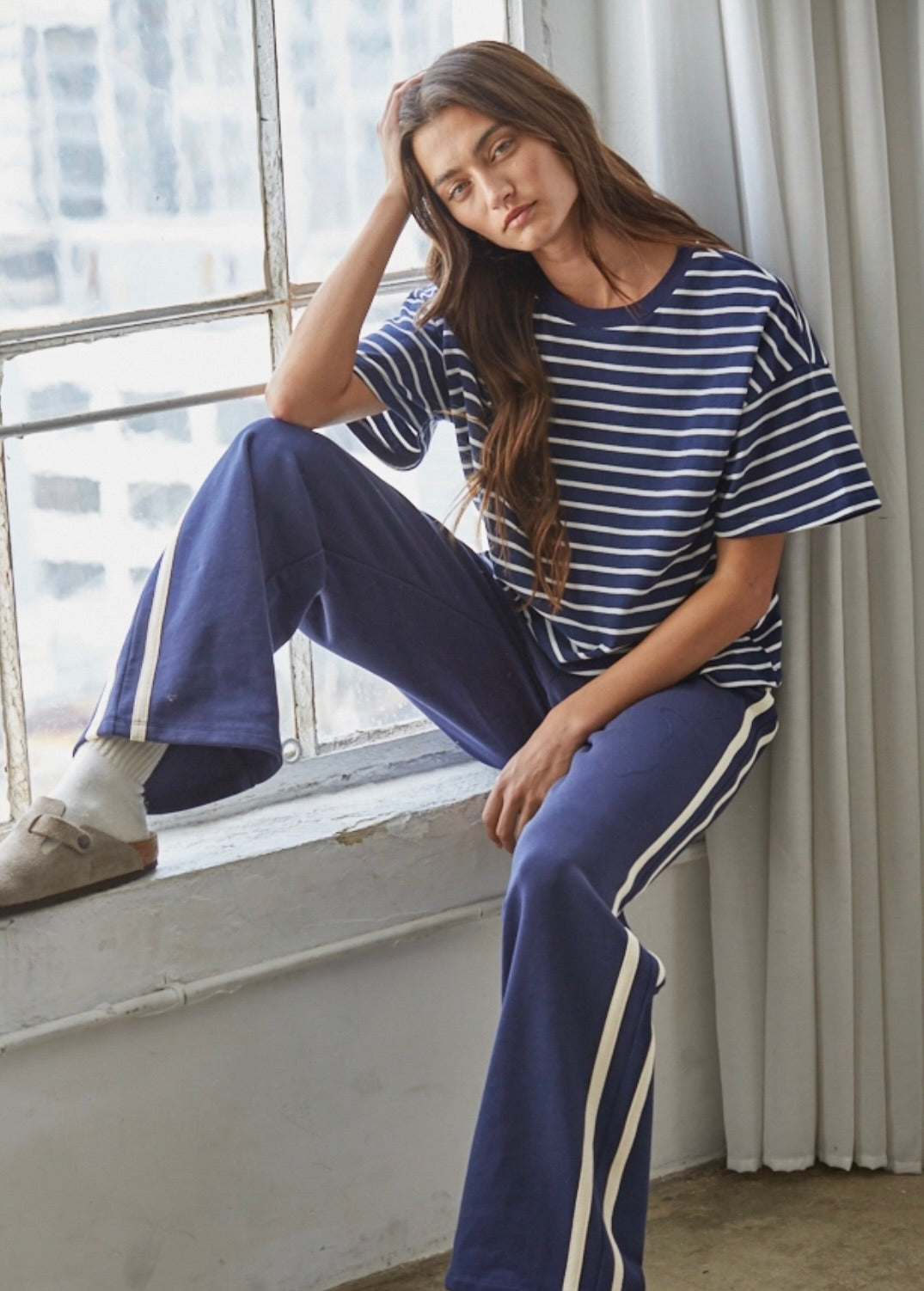 A woman sitting on a window sill wearing a navy blue and white striped short-sleeve t-shirt and matching navy blue wide-leg sweatpants with white side stripes.