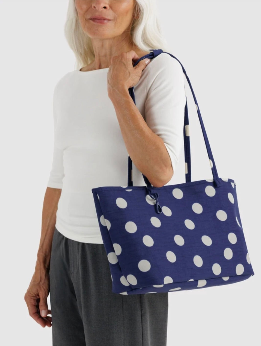 A woman wearing a white top and gray pants carries a navy blue tote bag with large white polka dots against a light gray studio background.