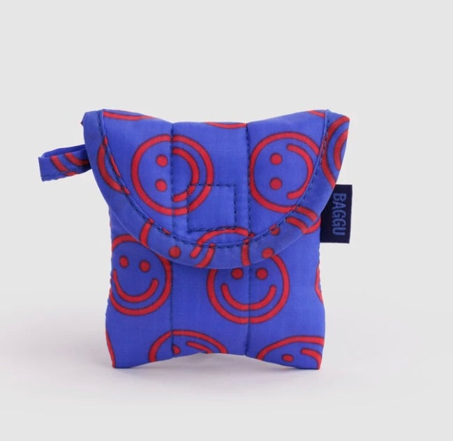 A small quilted pouch in bright blue with a red smiley face pattern, featuring a top flap and a small black brand tag that reads BAGGU.
