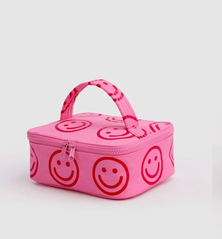 A pink zippered lunch bag with a top handle, featuring an all-over red smiley face pattern, photographed on a light gray background.