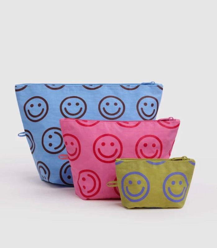 Three fabric zip pouches in blue, pink, and olive green, each printed with a repeating smiley face pattern in contrasting colors, arranged on a light gray background.