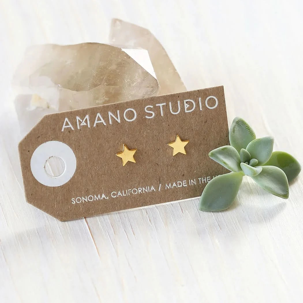 Gold star earrings on a 'Amano Studio' branded card with a succulent plant on a light wooden surface.