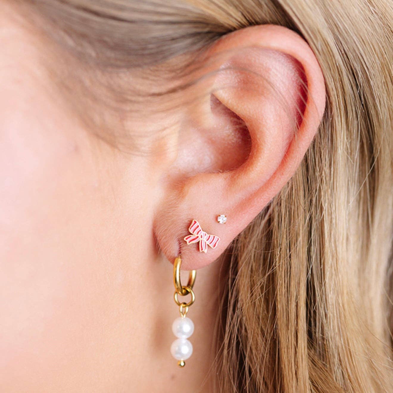 Close-up of an ear wearing a gold hoop earring with pearl drop and a small bow-shaped earring.