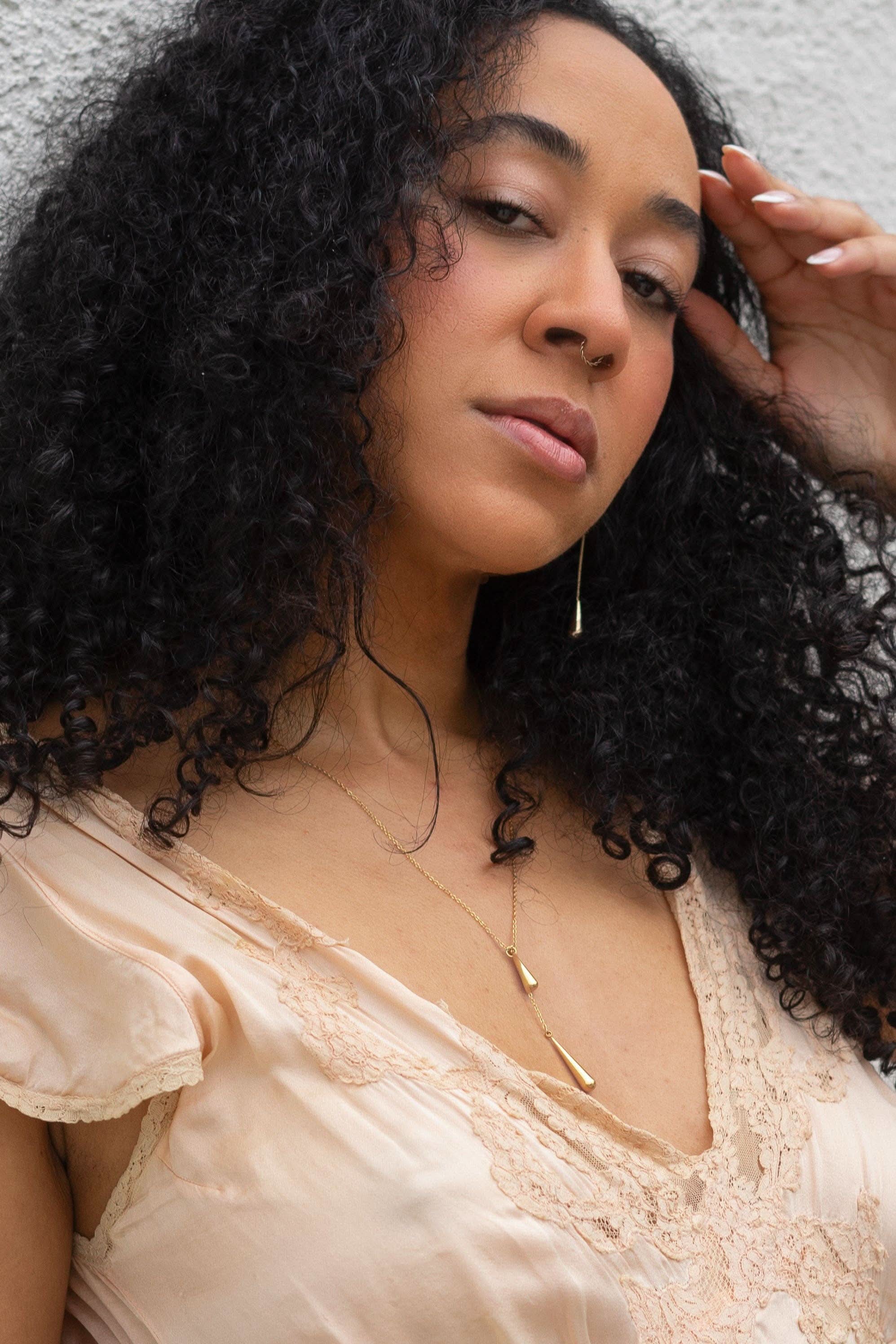 Woman with curly hair wearing a beige top and gold necklace against a neutral background