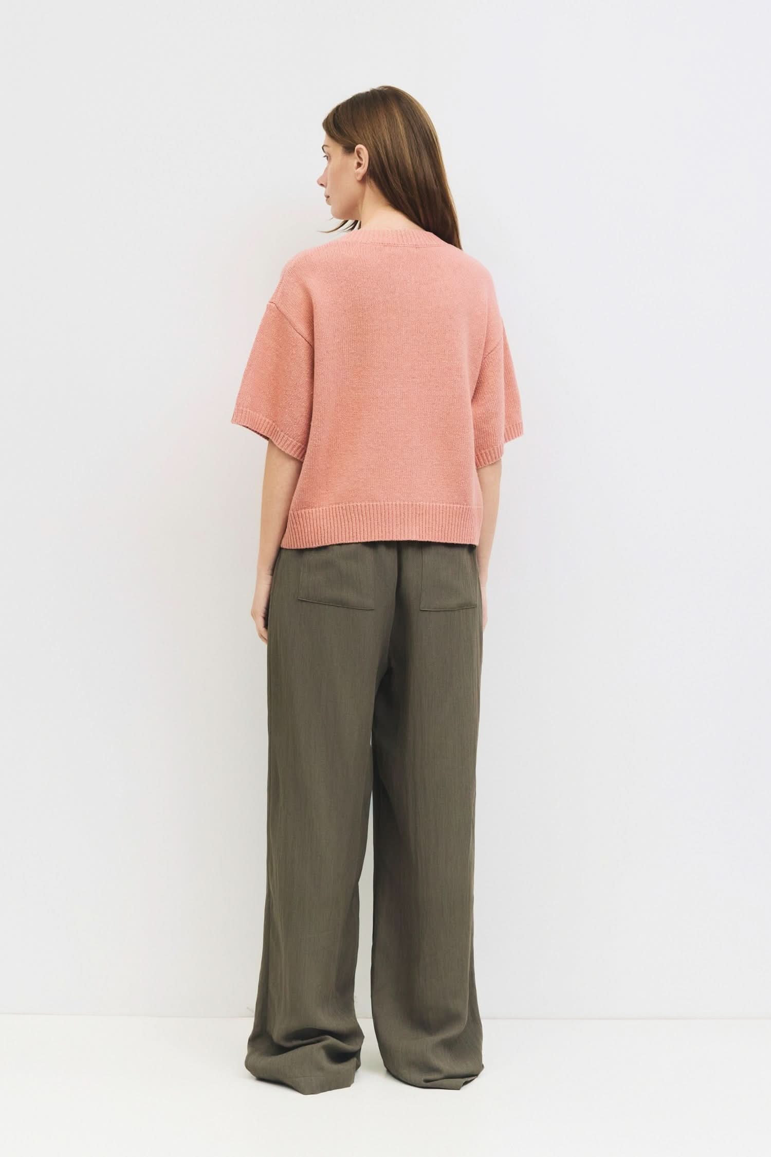 Woman standing against a white wall wearing a salmon pink, short sleeved, sweater cardigan 