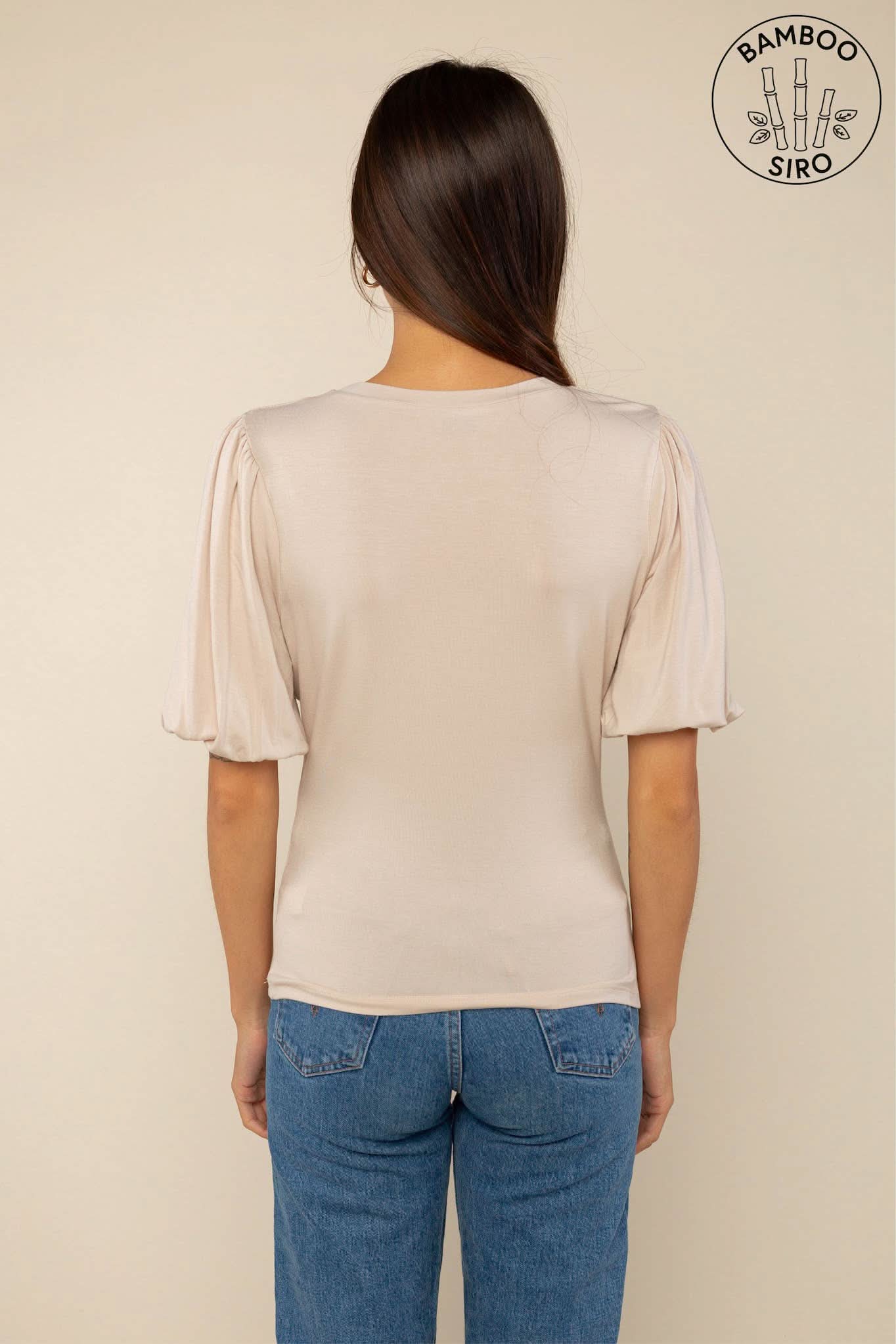 Woman wearing a sand color crew neck top with puffy sleeves and blue jeans with back pockets behind a blank background
