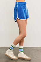 Woman wearing tube socks, tennis shoes, and electric marine blue terry cloth towel shorts with white lining details and white drawstring with hand in pocket 
