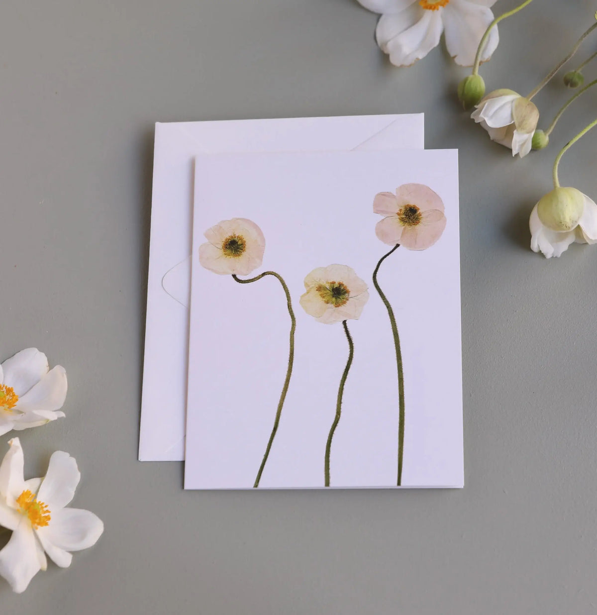 A white greeting card with a pressed floral design of three pale pink poppy-like flowers and long green stems, shown with a matching white envelope on a gray surface surrounded by white flowers.