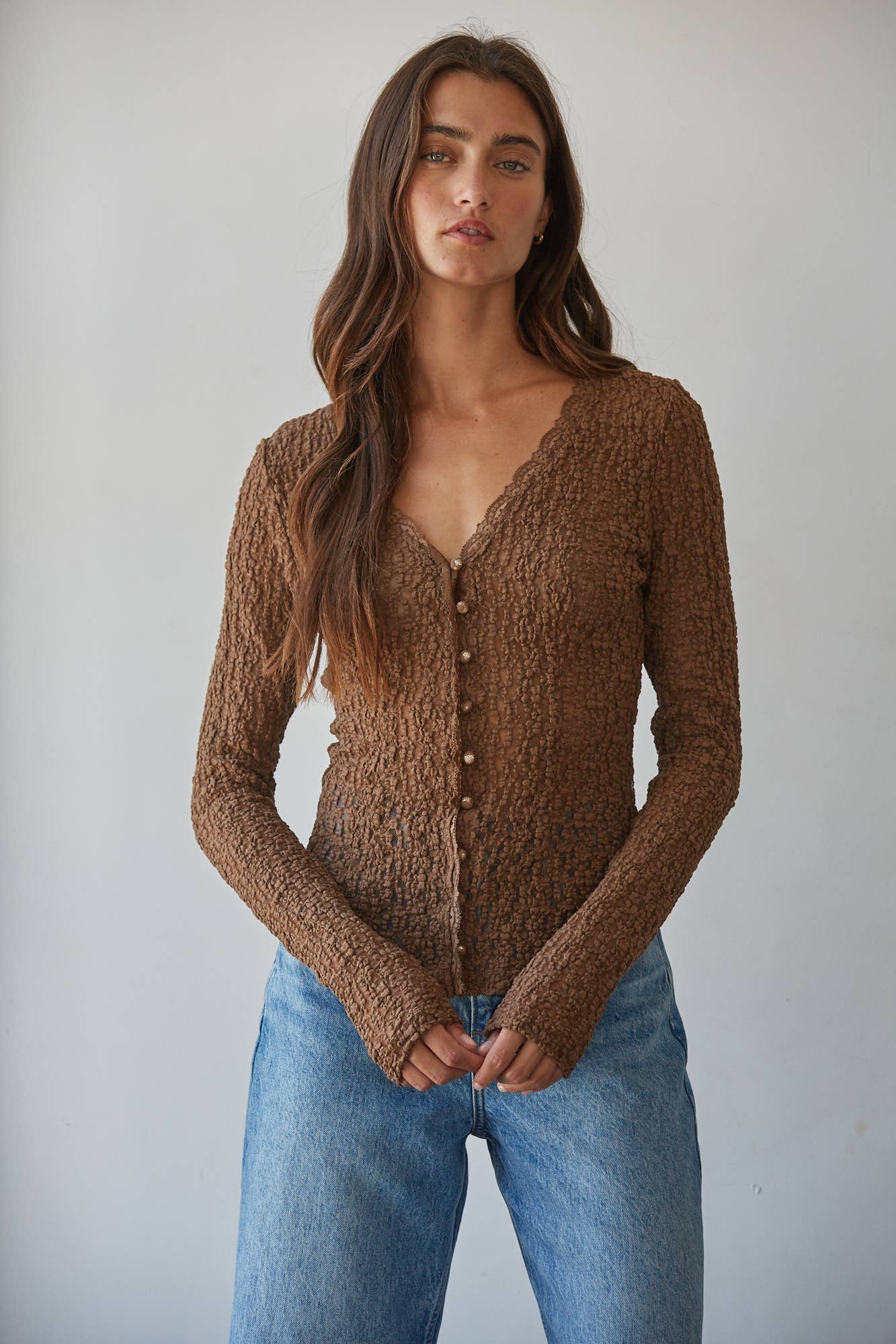 A woman wearing a brown textured knit cardigan with a scalloped V-neck and button-front closure, paired with light blue jeans, photographed against a plain light gray studio background.