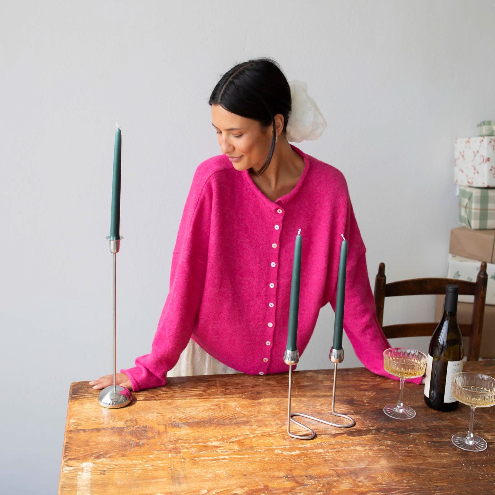 Woman smiling standing against white background leaning on wooden table arranging green taper candles wearing Fuschia pinkish button down cardigan with white buttons and white skirt
