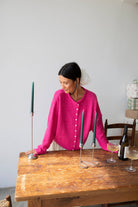 Woman smiling standing against white background leaning on wooden table arranging green taper candles wearing Fuschia pinkish button down cardigan with white buttons and white skirt
