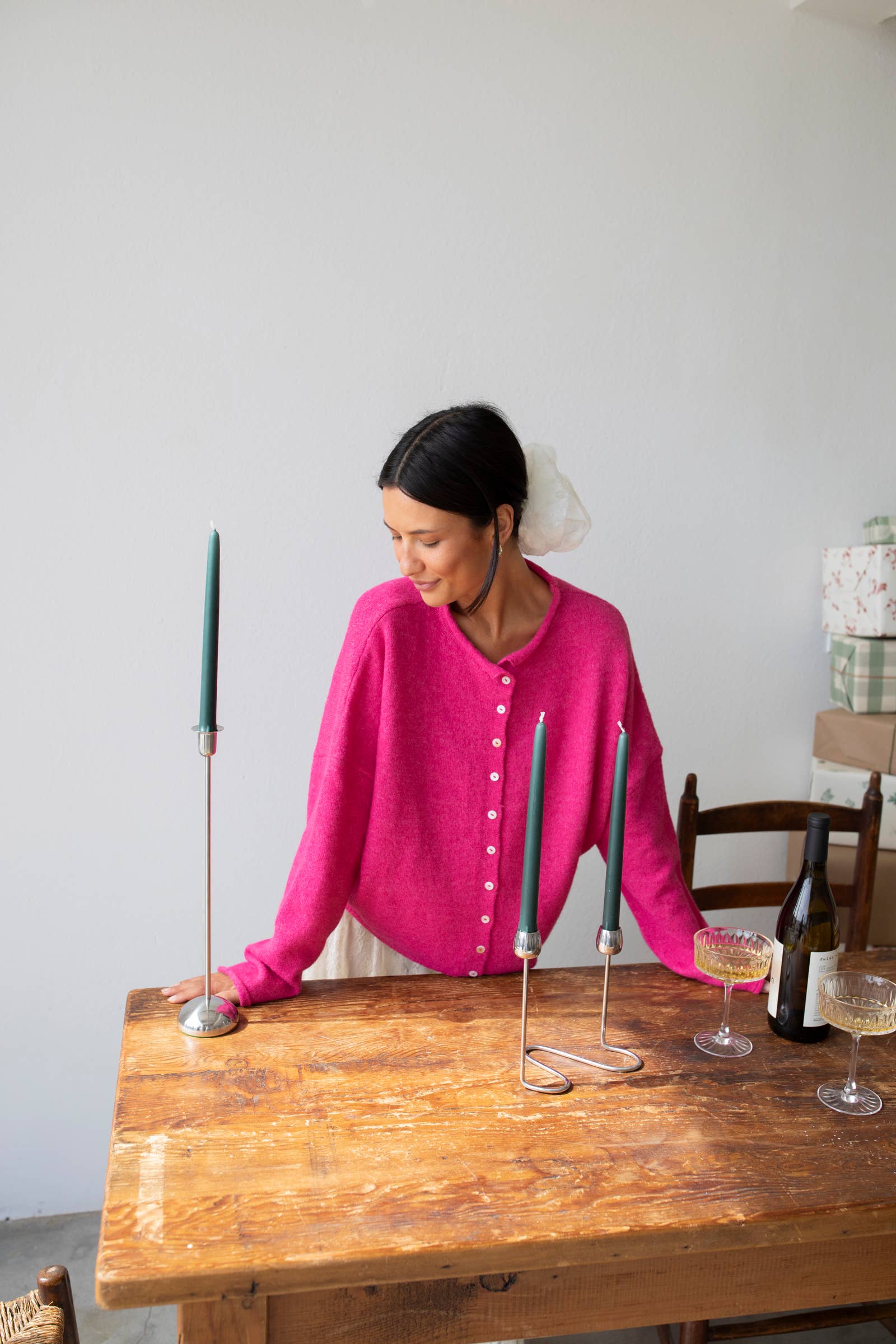 Woman smiling standing against white background leaning on wooden table arranging green taper candles wearing Fuschia pinkish button down cardigan with white buttons and white skirt