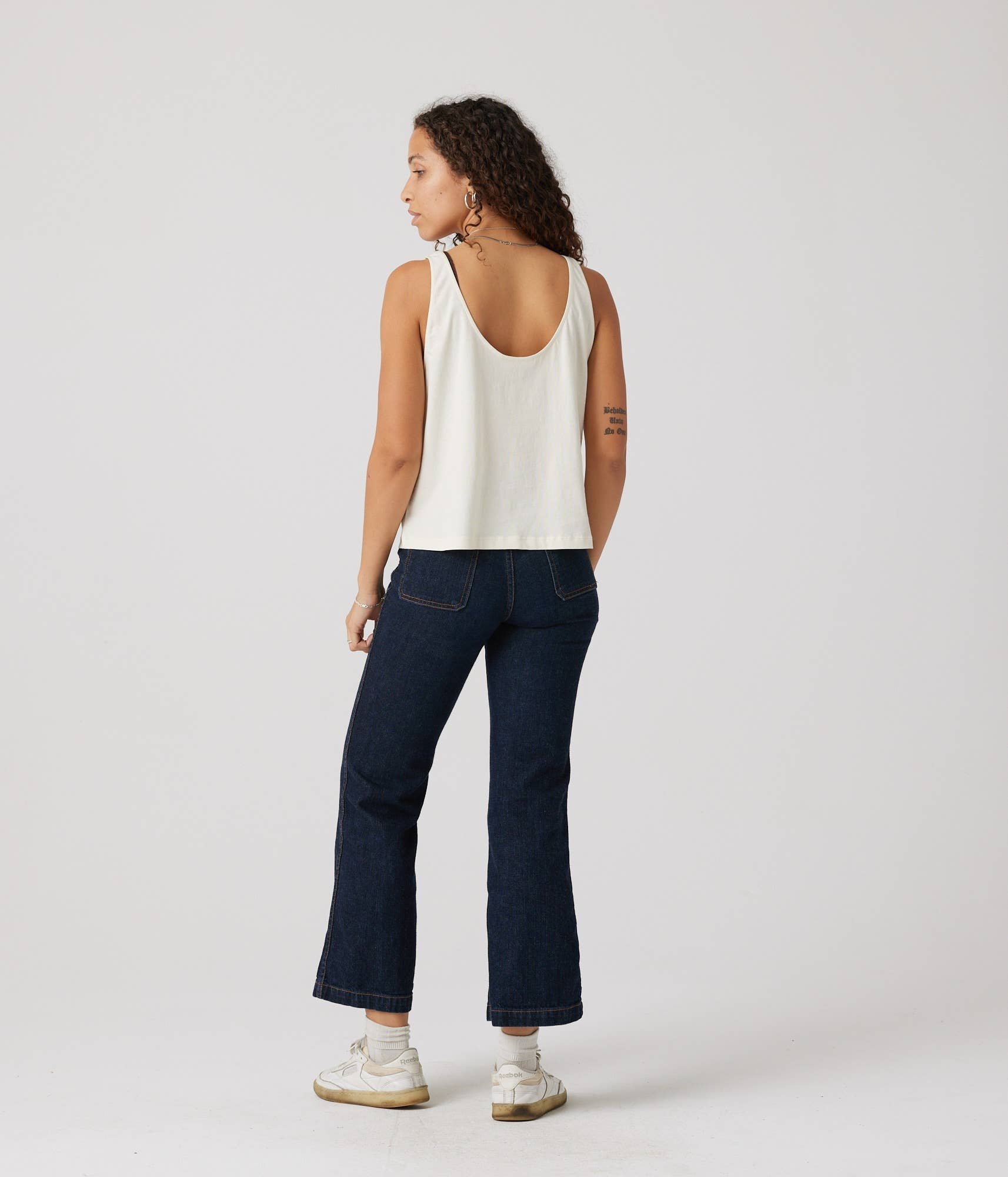 Person wearing a white tank top and dark blue jeans on a plain background