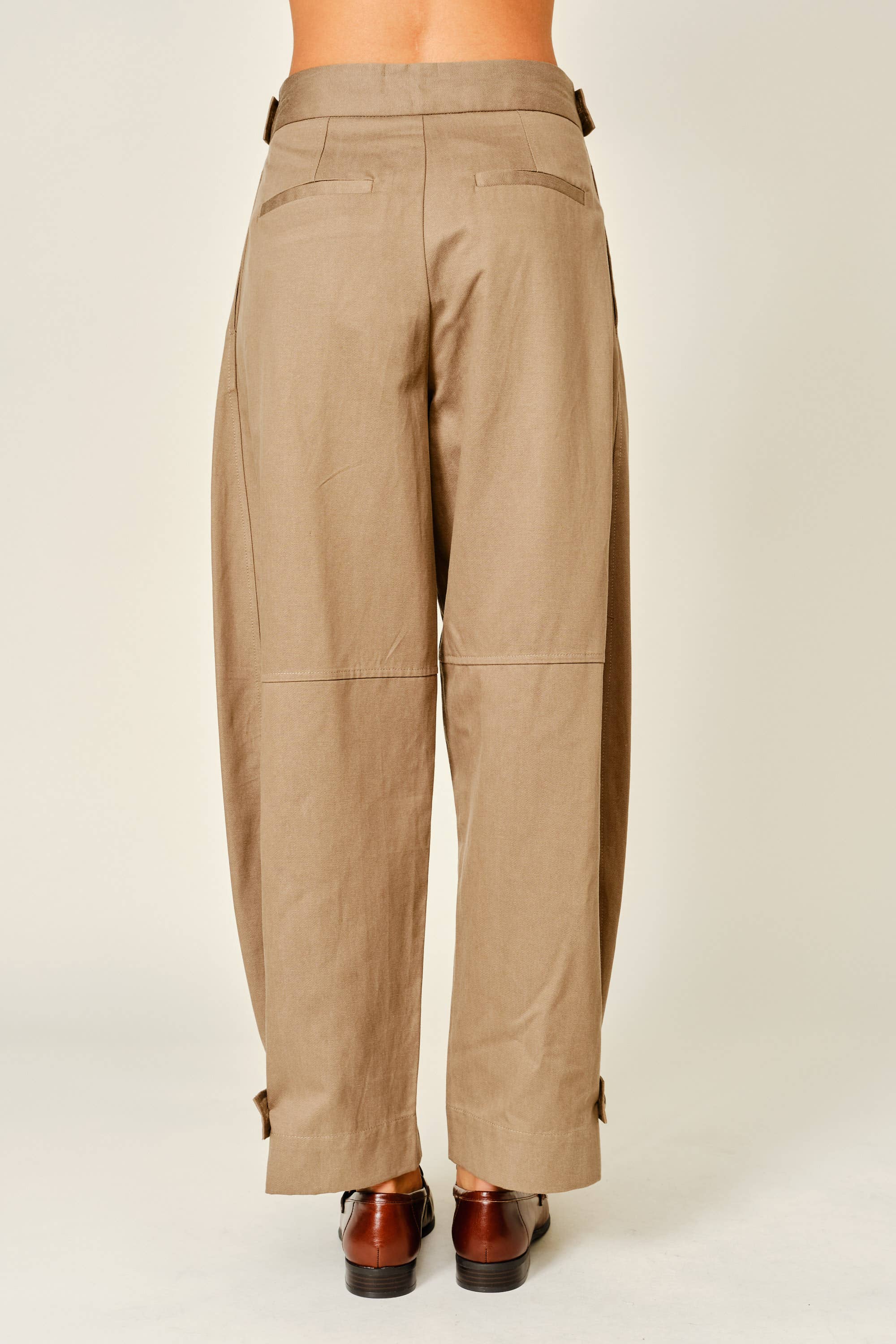 A person shown from the back wearing loose, wide-leg tan pants with belt loops, flap back pockets, and cuffed hems, paired with brown shoes against a plain light studio background.