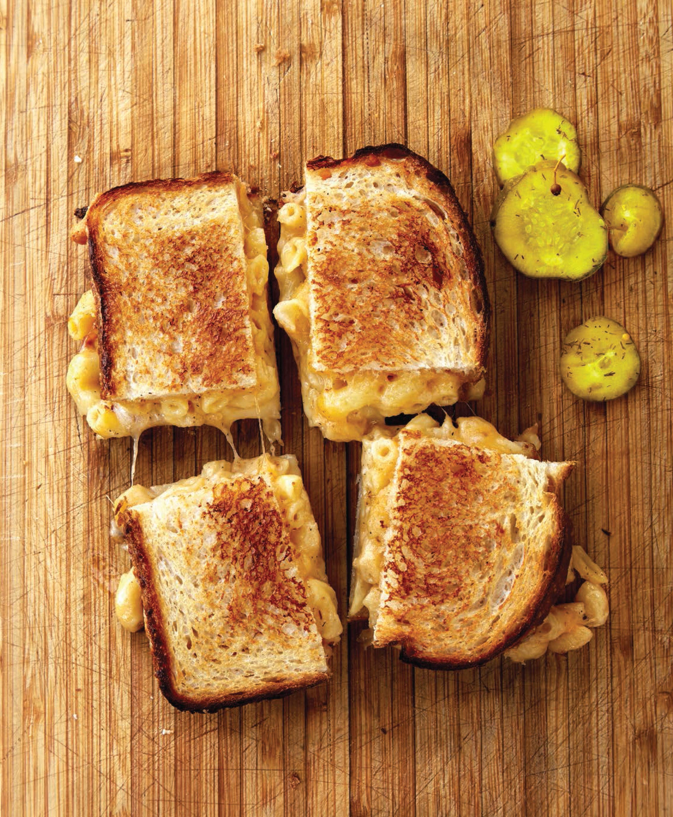 Four grilled cheese sandwiches on a wooden cutting board with pickles.