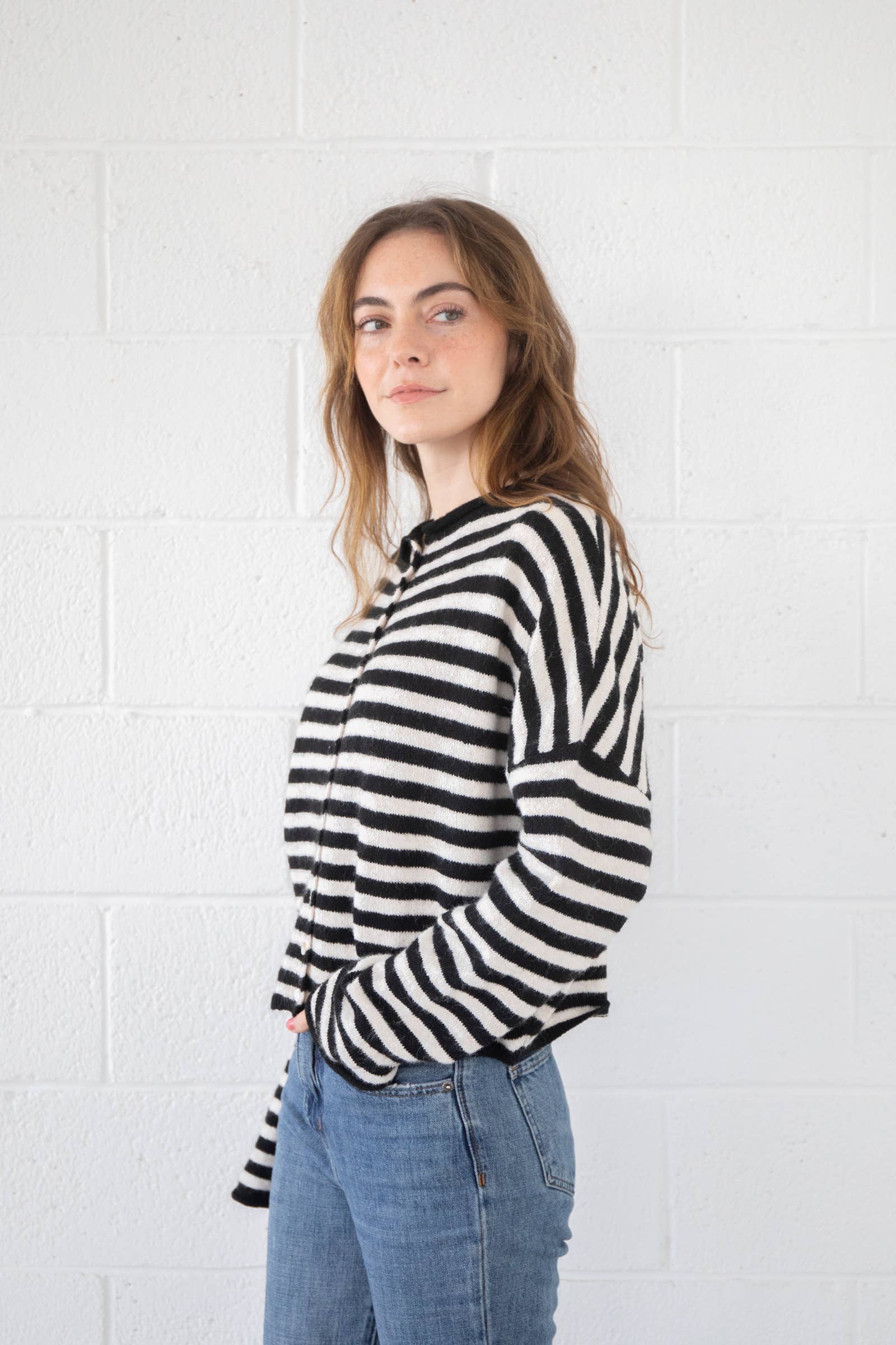 Woman wearing blue jeans and button down, black and white striped, cardigan standing against white brick background 