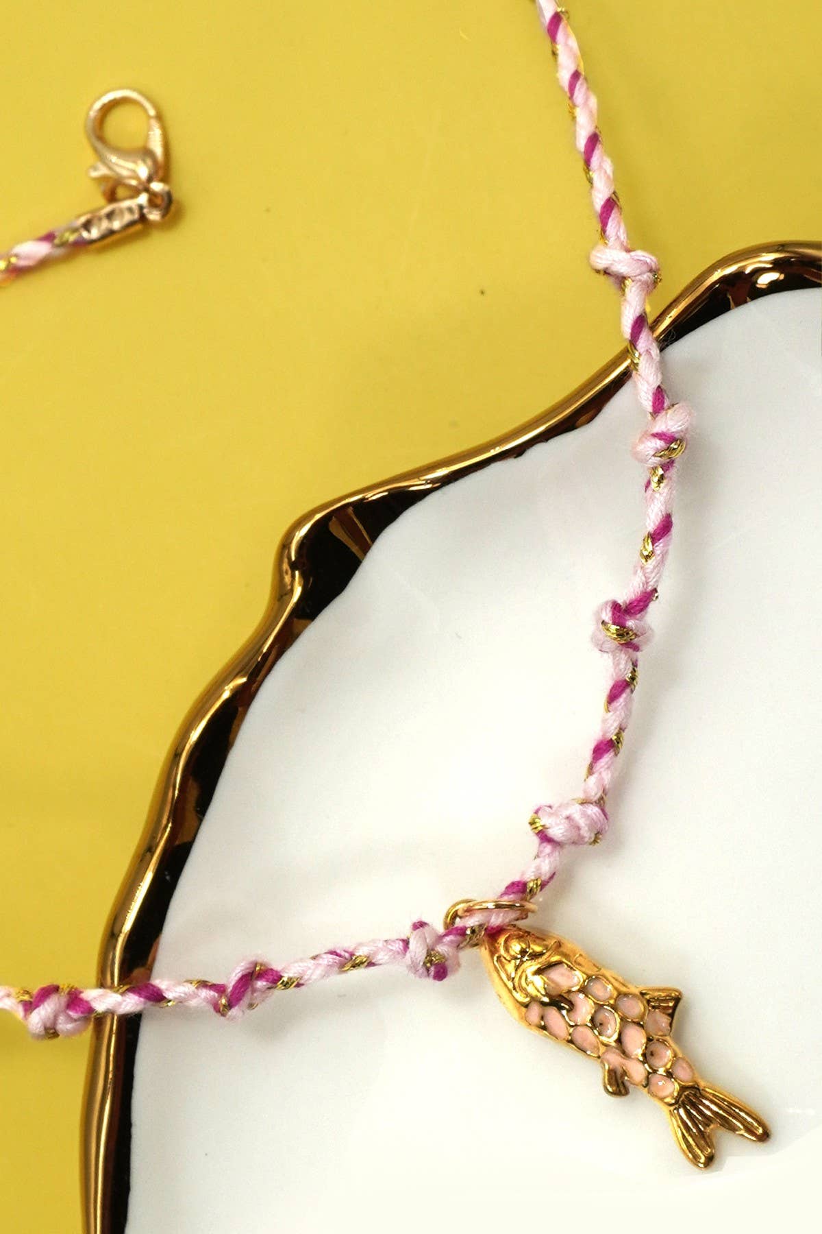 Pink braided bracelet with a gold fish charm on a white dish against a yellow background