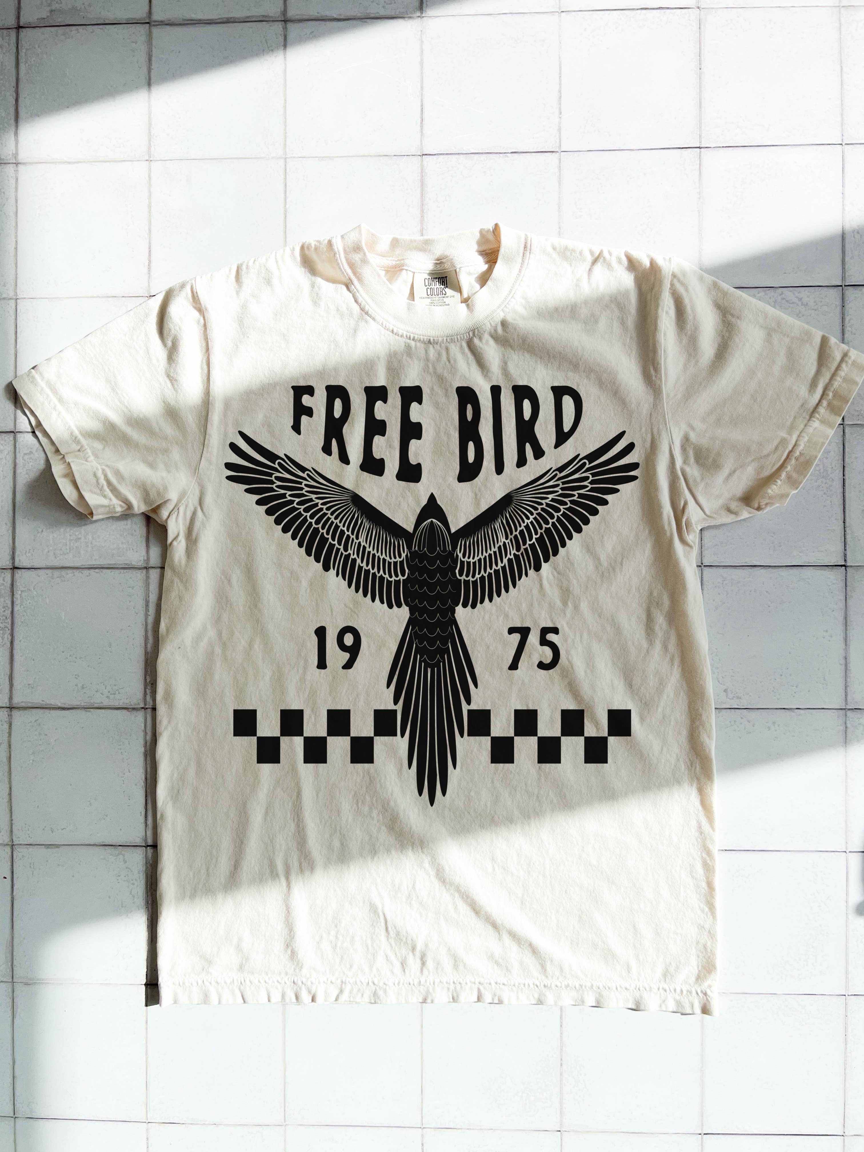 White t-shirt with black eagle graphic and text 'FREE BIRD 1975' on a tiled wall background