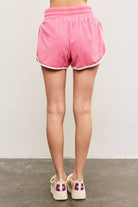 Woman facing away from camera wearing colorful crochet shoes and candy pink terry cloth towel shorts with white lining details and white drawstring with hand in pocket 