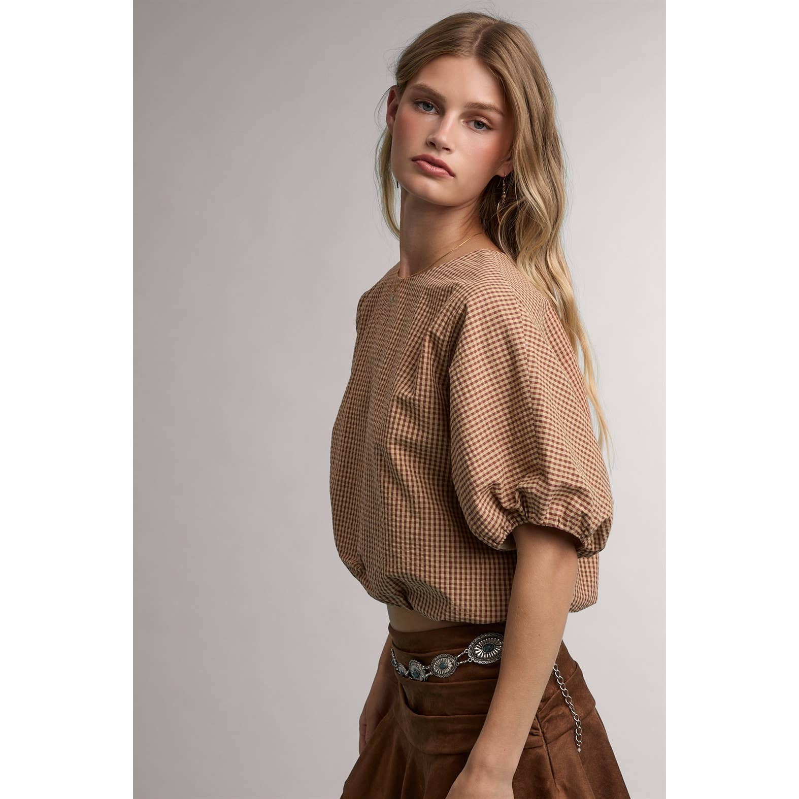 A woman modeling a brown and beige checkered puff-sleeve top with a cropped silhouette, paired with a brown skirt and a silver chain belt, against a plain gray studio background.