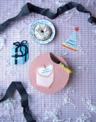 Light pink and red heart shaped happy birthday cake card on a purple plaid table surrounded by party items. 