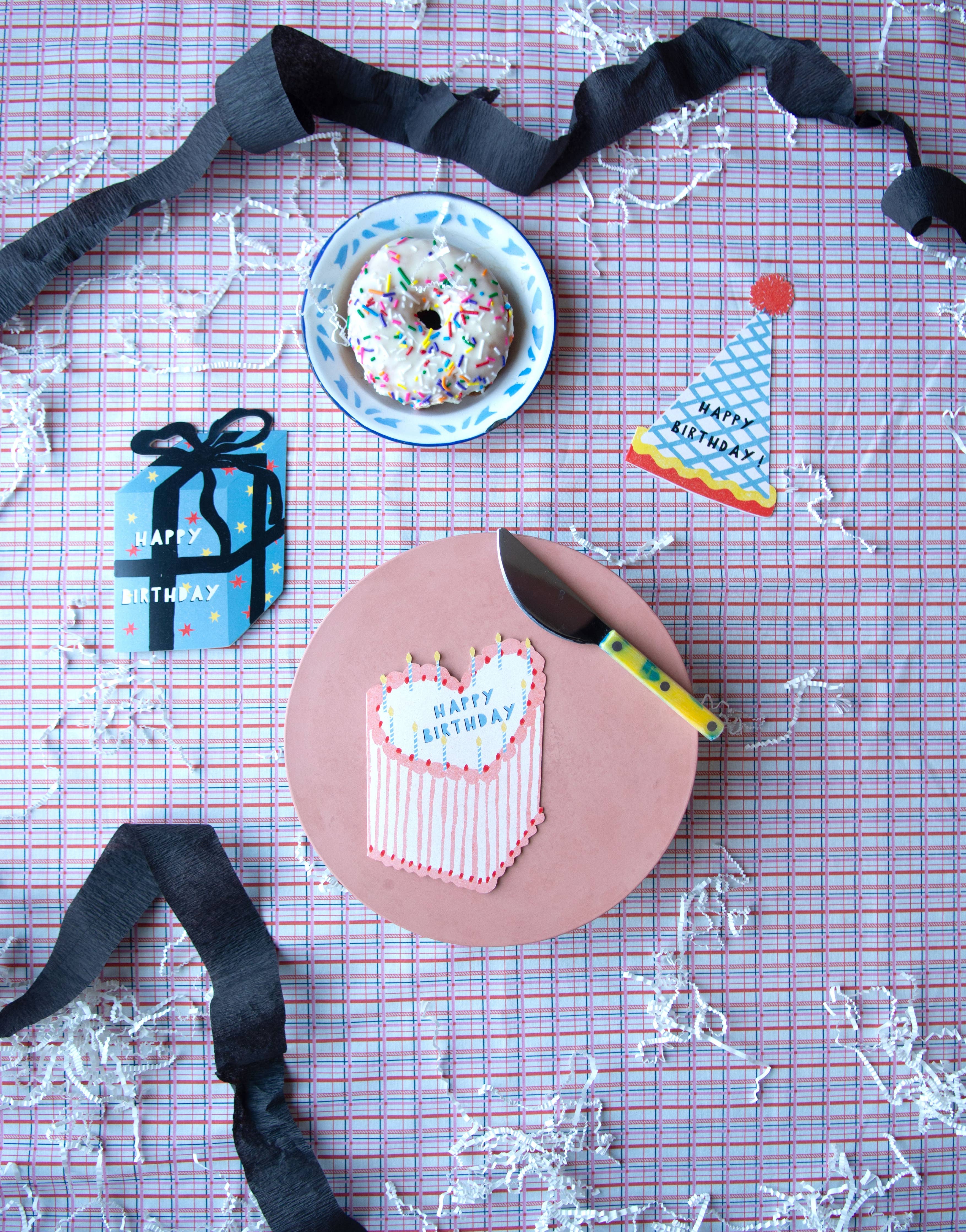 Light pink and red heart shaped happy birthday cake card on a purple plaid table surrounded by party items. 
