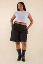 Woman wearing a grey sleeveless top with black vertical pin striped knee length shorts with pockets and black boots behind a blank background. 