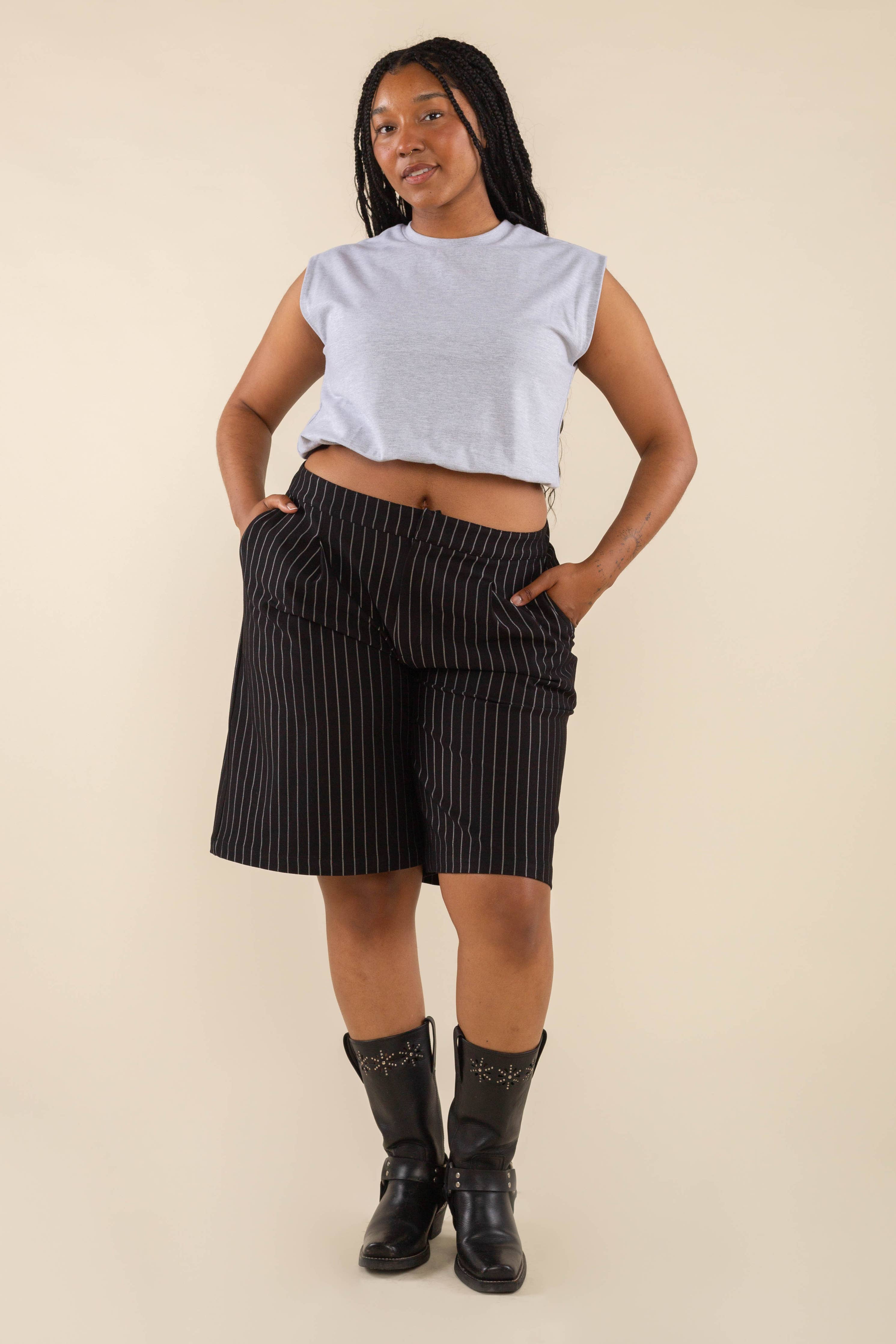 Woman wearing a grey sleeveless top with black vertical pin striped knee length shorts with pockets and black boots behind a blank background. 