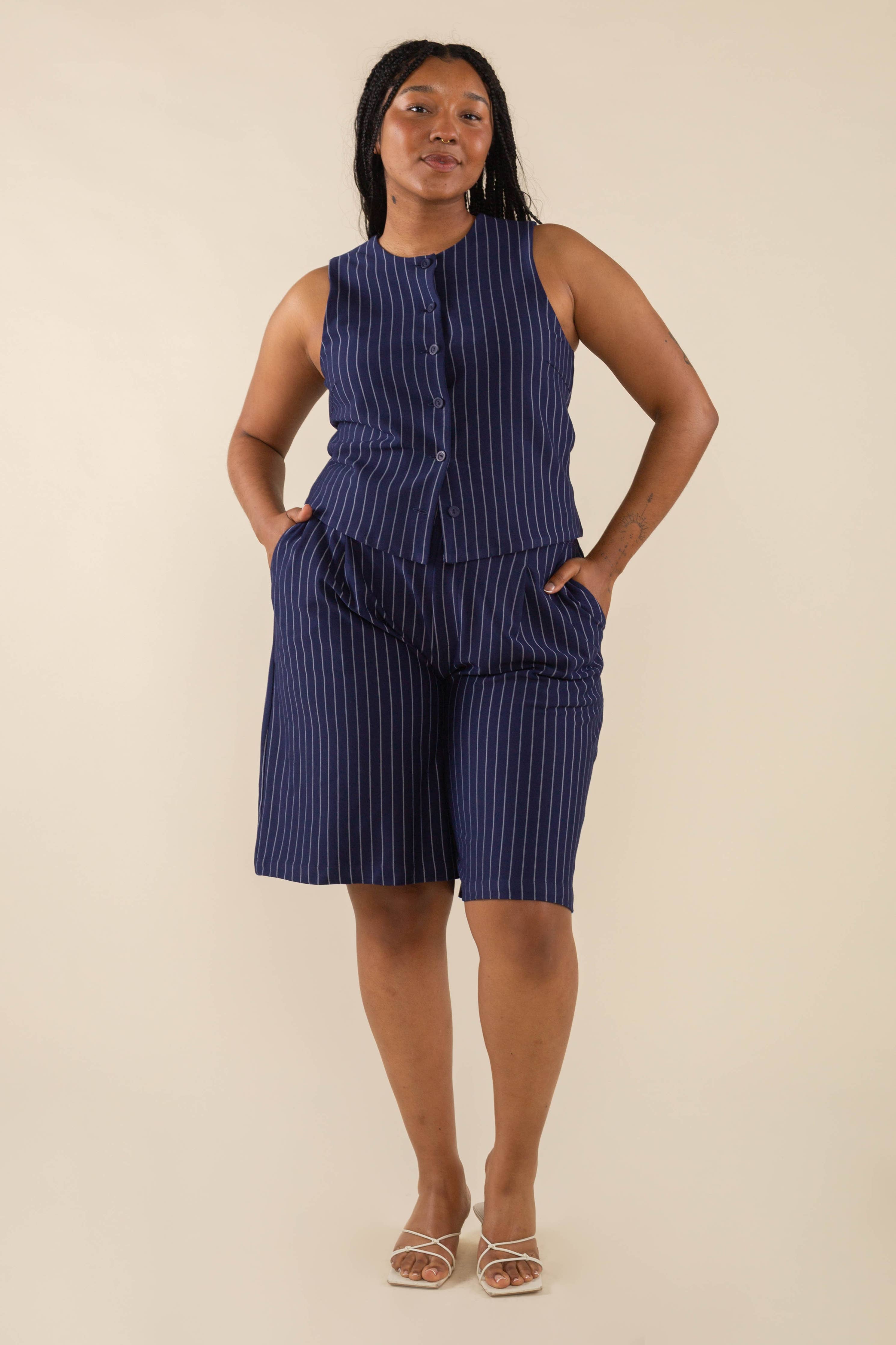 Woman wearing a navy vertical pin striped button down sleeveless top with navy vertical pin striped knee length shorts with pockets behind a blank background. 