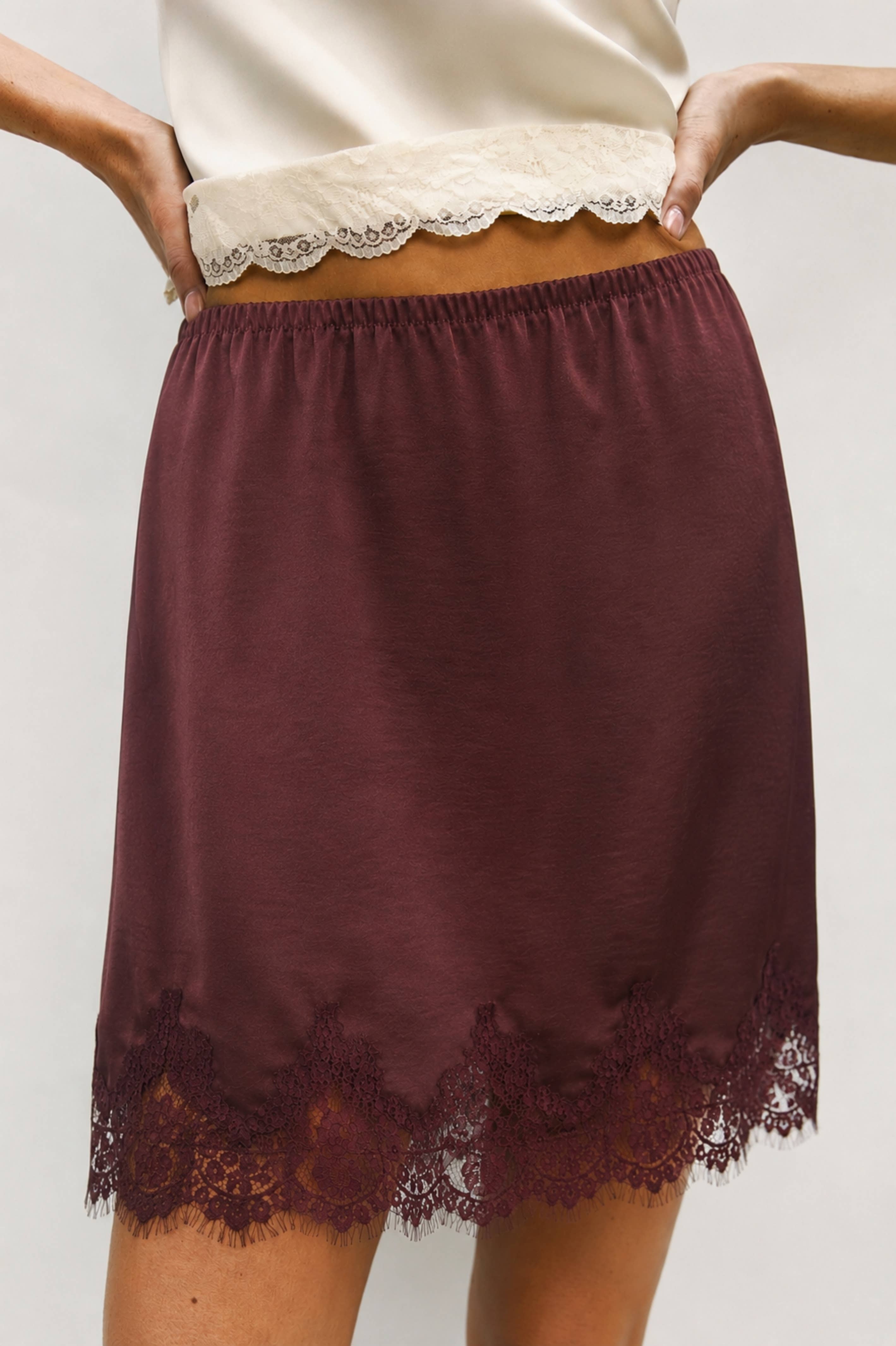 A close-up of a model wearing a dark burgundy slip skirt with a lace hem and a matching lace waistband.