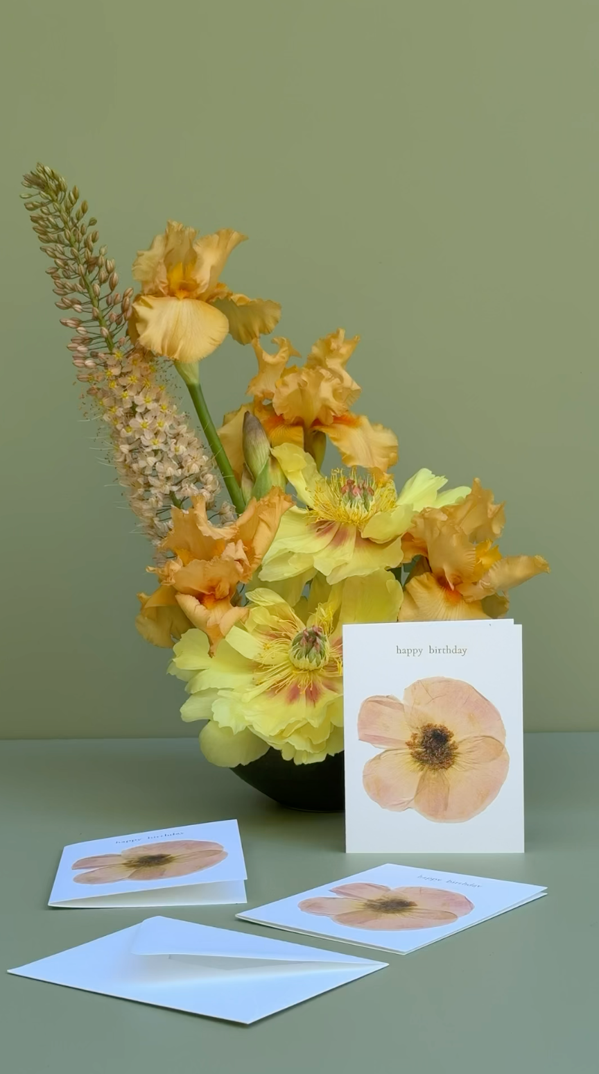 A floral arrangement with yellow and peach flowers in a dark vase on a muted green surface, alongside several white greeting cards featuring a peach poppy illustration and envelopes.