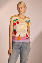 Woman wearing sleeveless, ivory, colorful, crochet, top with cherries, lemon, strawberry, watermelon, pear, and pineapple with pink, purple, and yellow horizontal stripes 