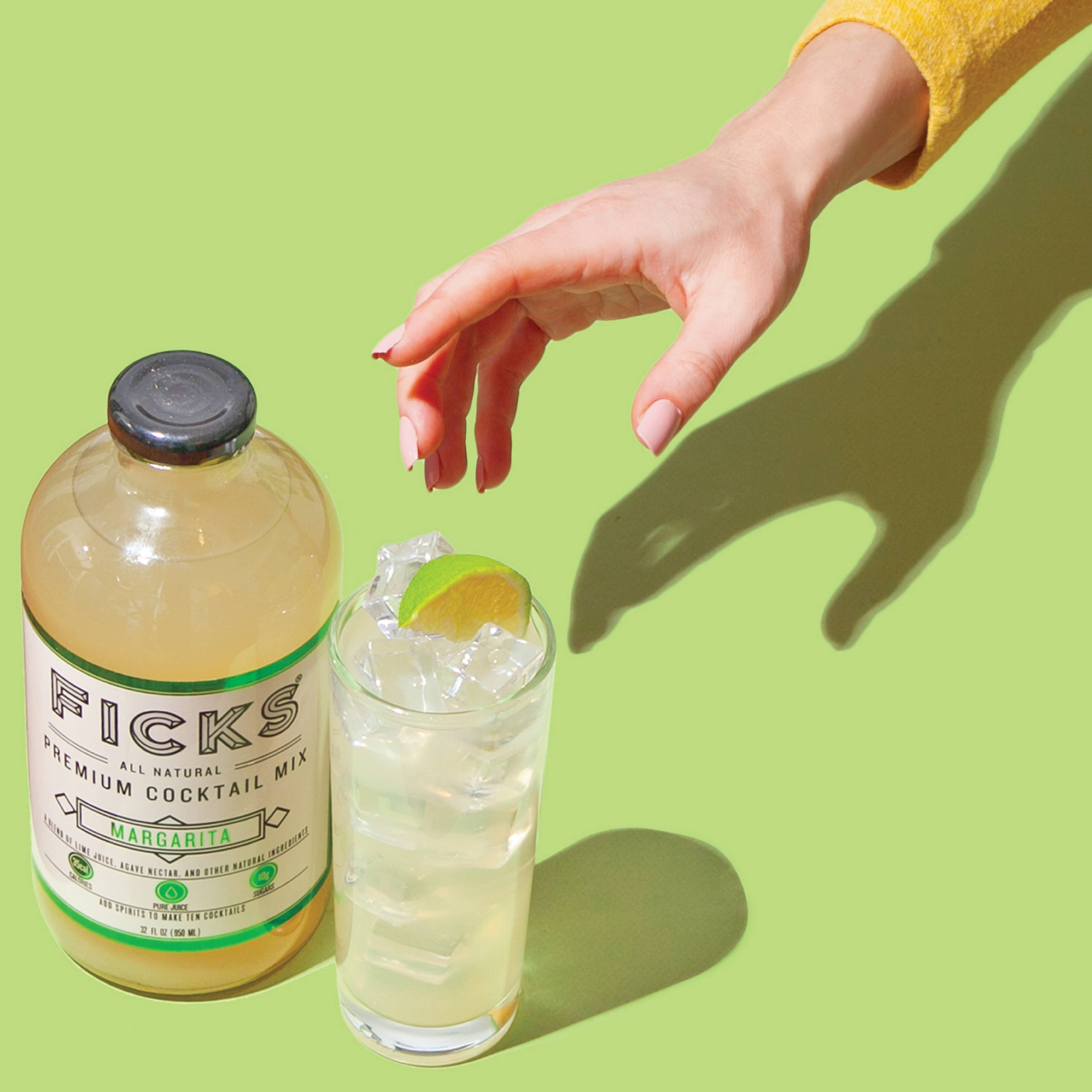 Glass cocktail bottle sitting on green surface next to drinking glass containing cocktail with hand reaching for glass