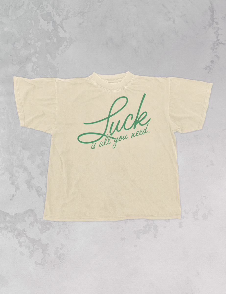 “Luck is all you need” written in green cursive on cream tee shirt playing on a gray marbled backgound