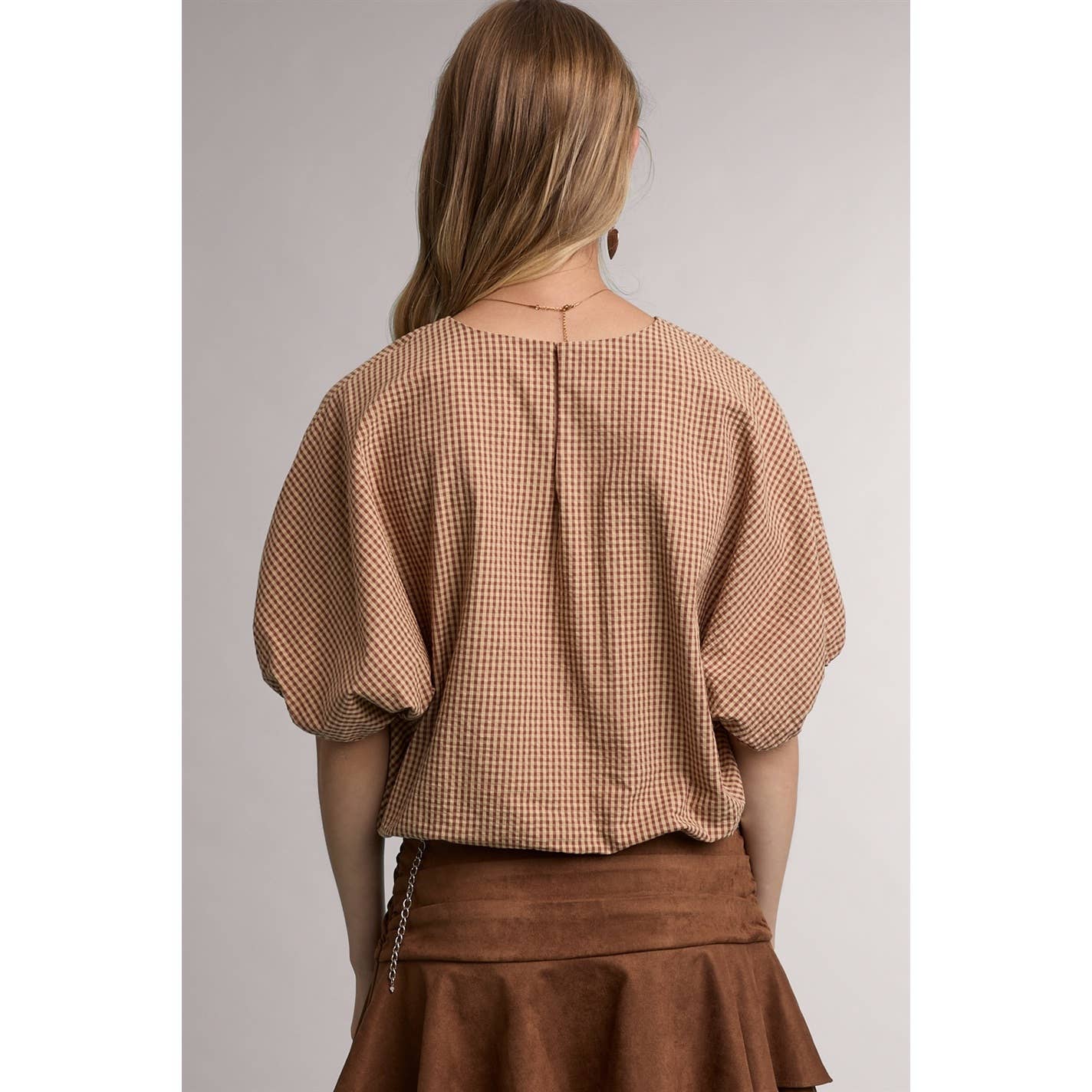 A woman shown from the back wearing a cropped short-sleeve blouse with a brown and beige gingham pattern and a brown skirt, set against a plain light gray studio background.