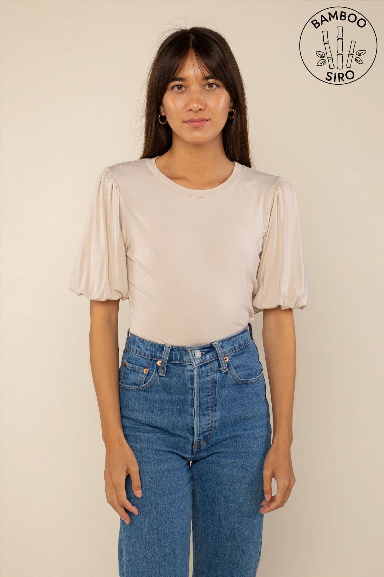 Woman wearing a sand color crew neck top with puffy sleeves and blue jeans behind a blank background. 
