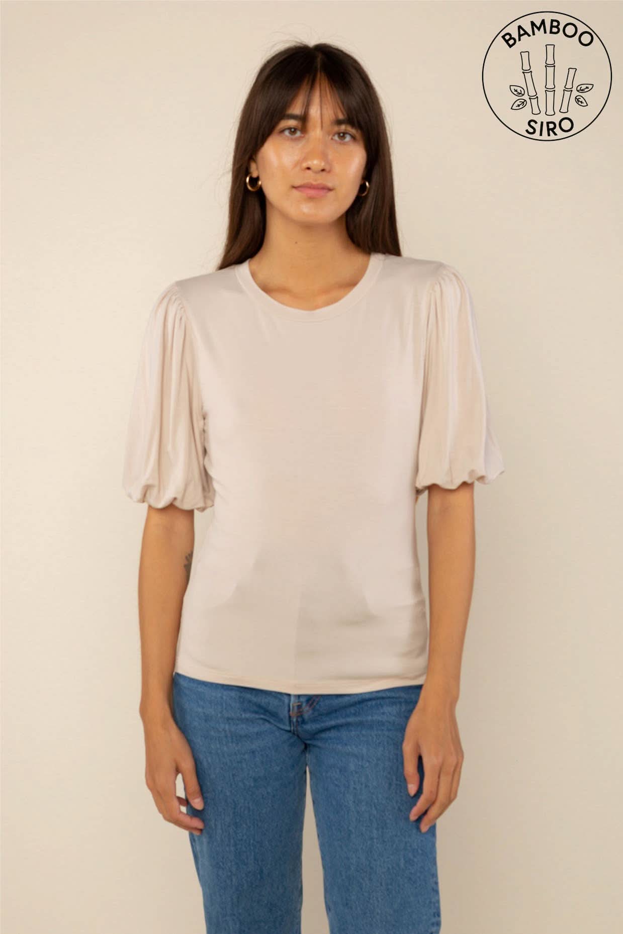 Woman wearing a sand color crew neck top with puffy sleeves and blue jeans behind a blank background