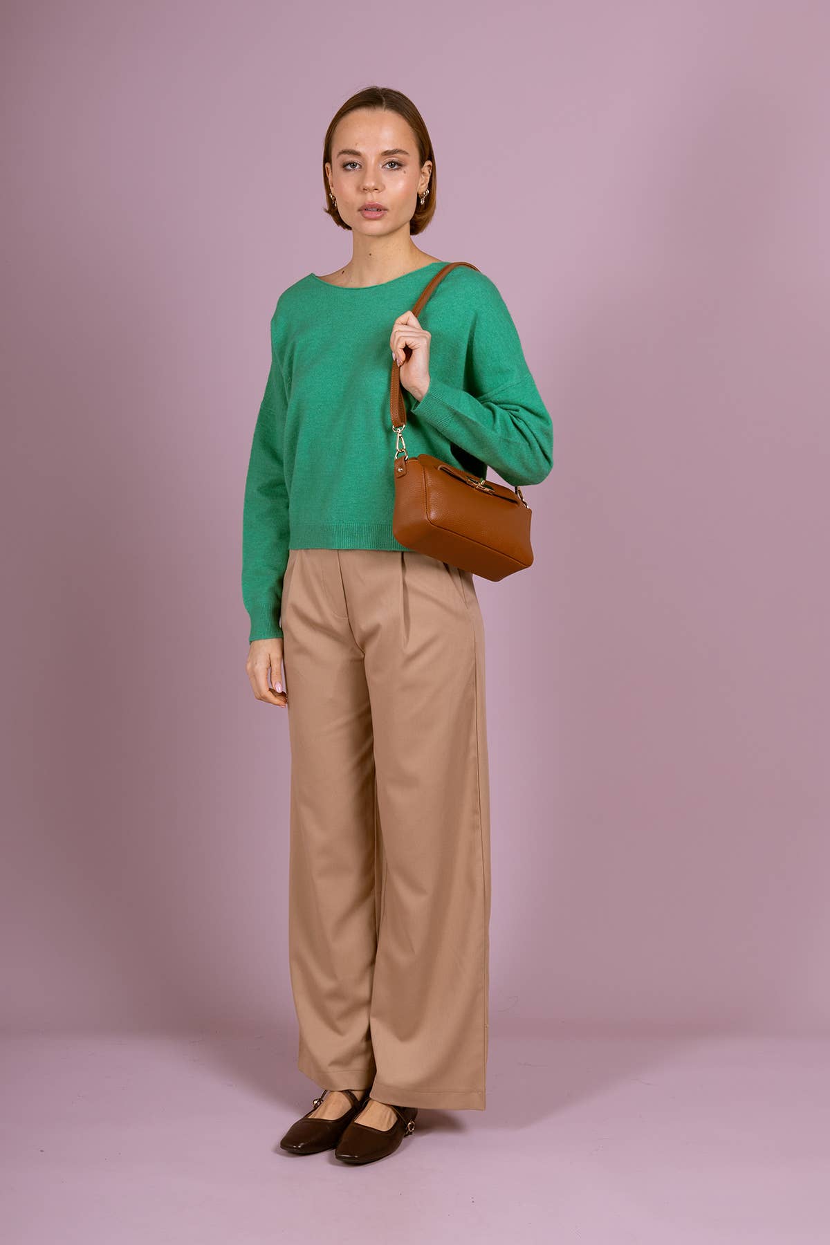 A woman stands against a solid lavender studio background wearing a vibrant green long-sleeve knit sweater, beige wide-leg trousers, and dark brown flats. She is holding a brown leather shoulder bag.