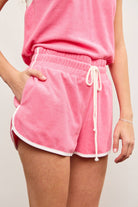 Woman wearing  candy pink terry cloth towel shorts with white lining details and white drawstring with hand in pocket 