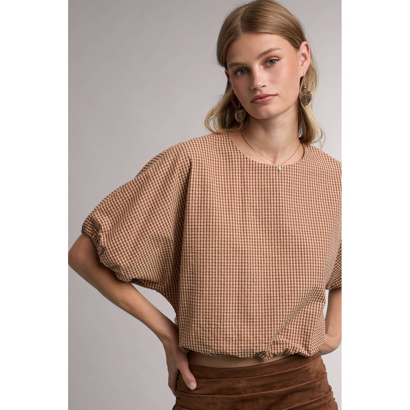 A woman wearing a brown and beige gingham short-sleeve top with voluminous puff sleeves, paired with a brown skirt, against a neutral gray studio background.