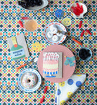 Yellow Happy Birthday cake with pink frosting and sprinkles card surrounded by food on a table with a colorful table cloth. 