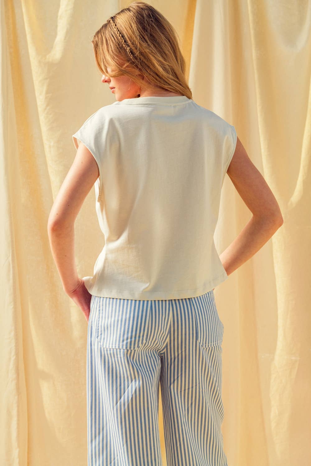 Person wearing a white sleeveless top and striped pants against a beige curtain.