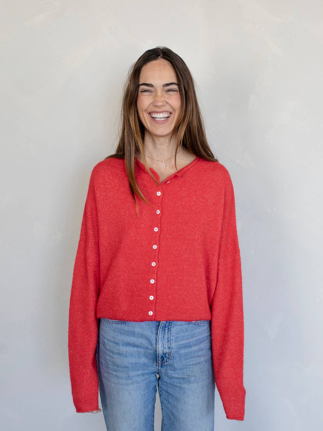 Woman smiling standing against white background wearing red button down cardigan with white buttons and blue jeans