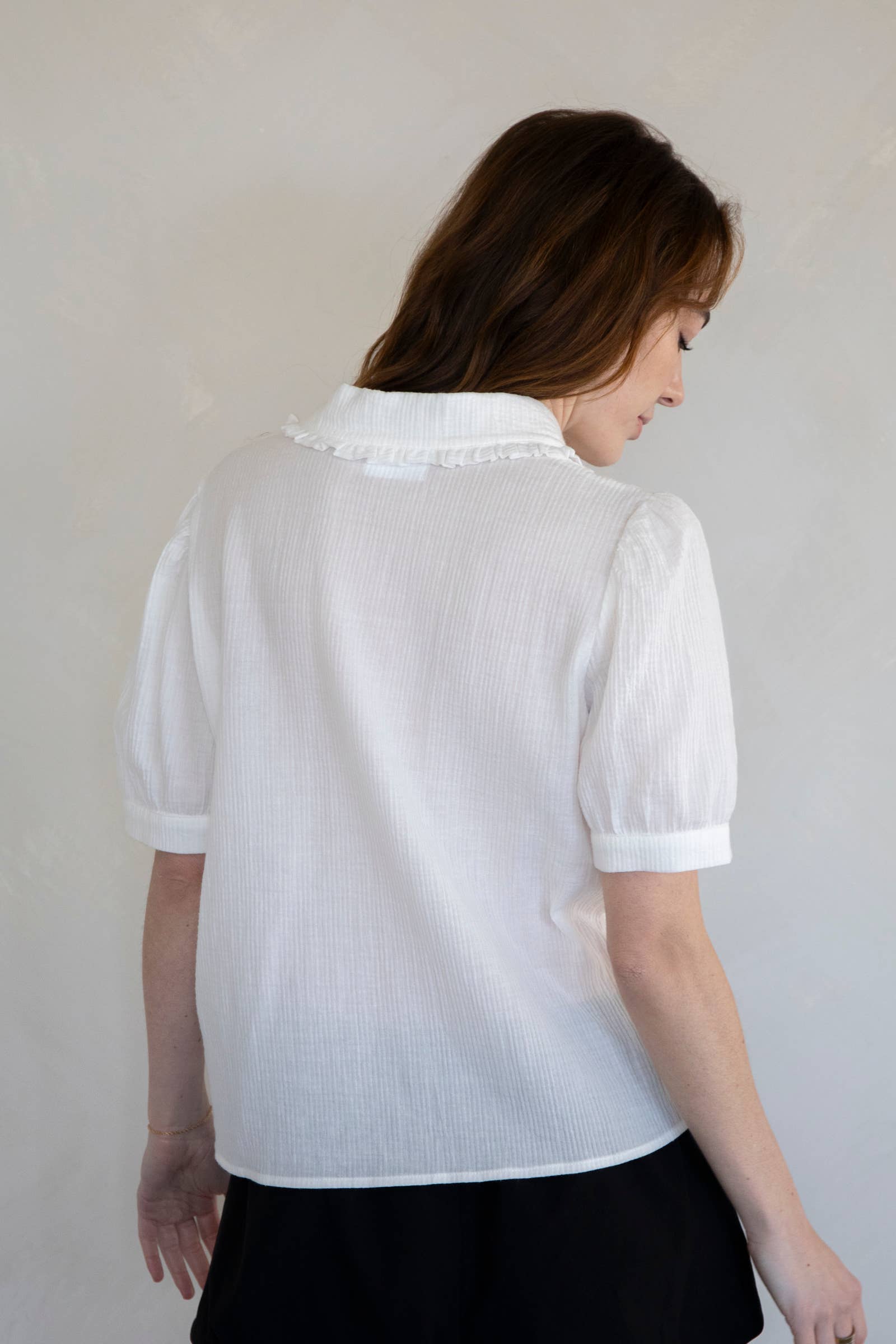 Woman standing facing away from the camera against white background wearing white collared button down blouse with extended sleeves and vertical line details 