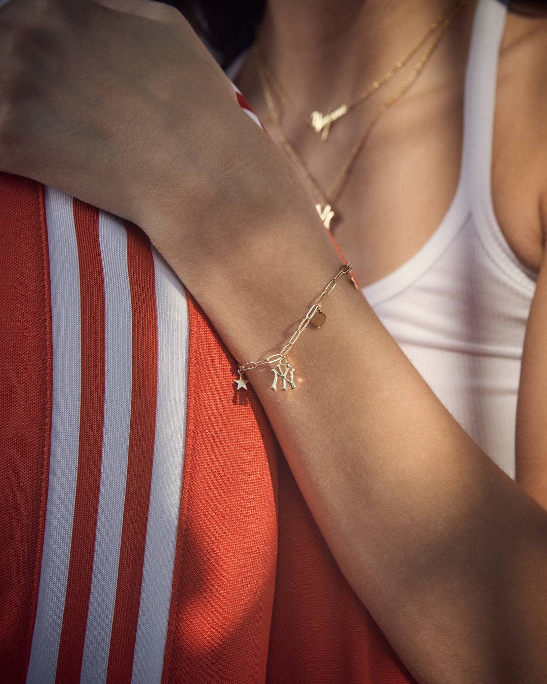A close-up lifestyle shot of a model wearing a delicate gold chain bracelet with small charms on her wrist, paired with a white tank top and an orange and white striped athletic jacket.