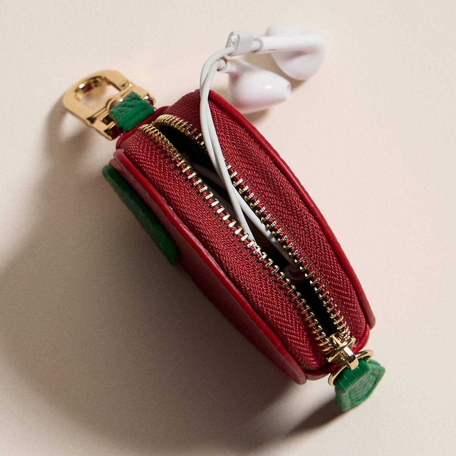 Red apple pouch with gold zipper and green accents containing white earbuds on a beige background